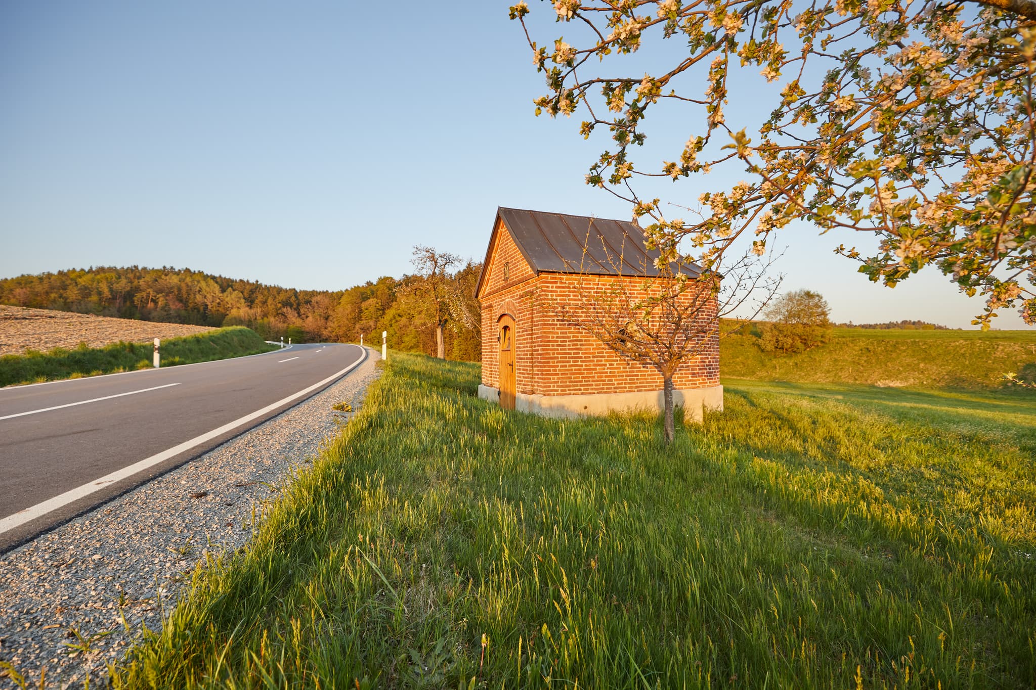 Kleine Backsteinkapelle im Abendlicht in Schildthurn bei Zeilarn, Landkreis Rottal-Inn, Niederbayern, Deutschland. Umgeben von grünen Wiesen und Bäumen.