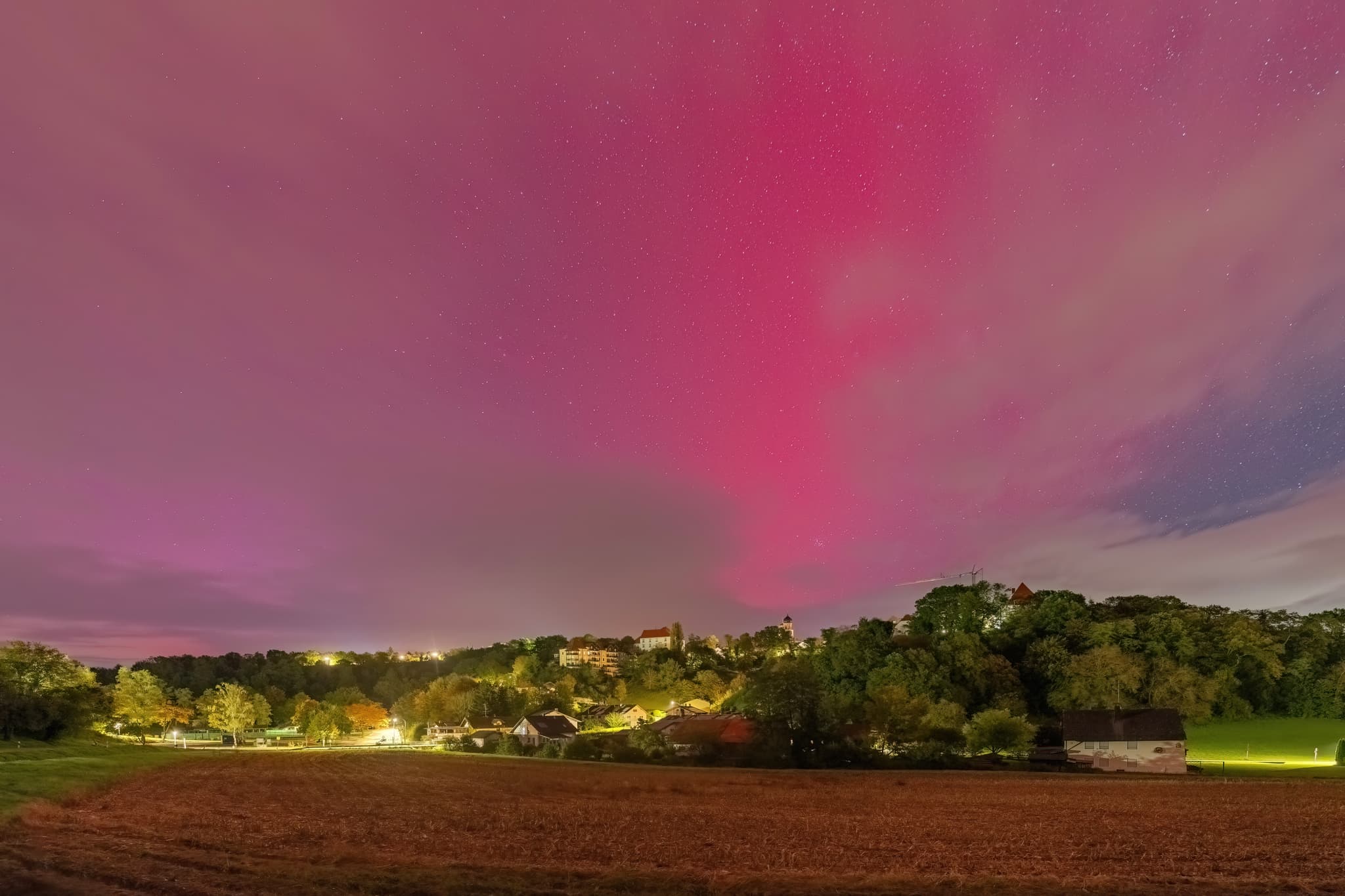Polarlichter am Himmel über Bad Griesbach, Stadt im Landkreis Passau, Niederbayern. Beleuchtete Siedlung mit Feldern in der Region Donau-Wald, Deutschland.