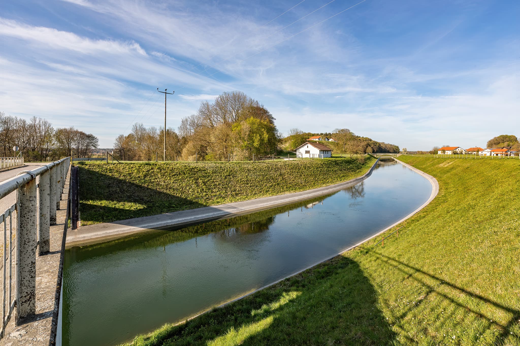 Der Alzkanal in Eschlberg, Mehring, Landkreis Altötting, Oberbayern. Landschaft der Inn-Salzach-Region in Deutschland mit Wasserlauf, grünen Ufern und Gebäuden.
