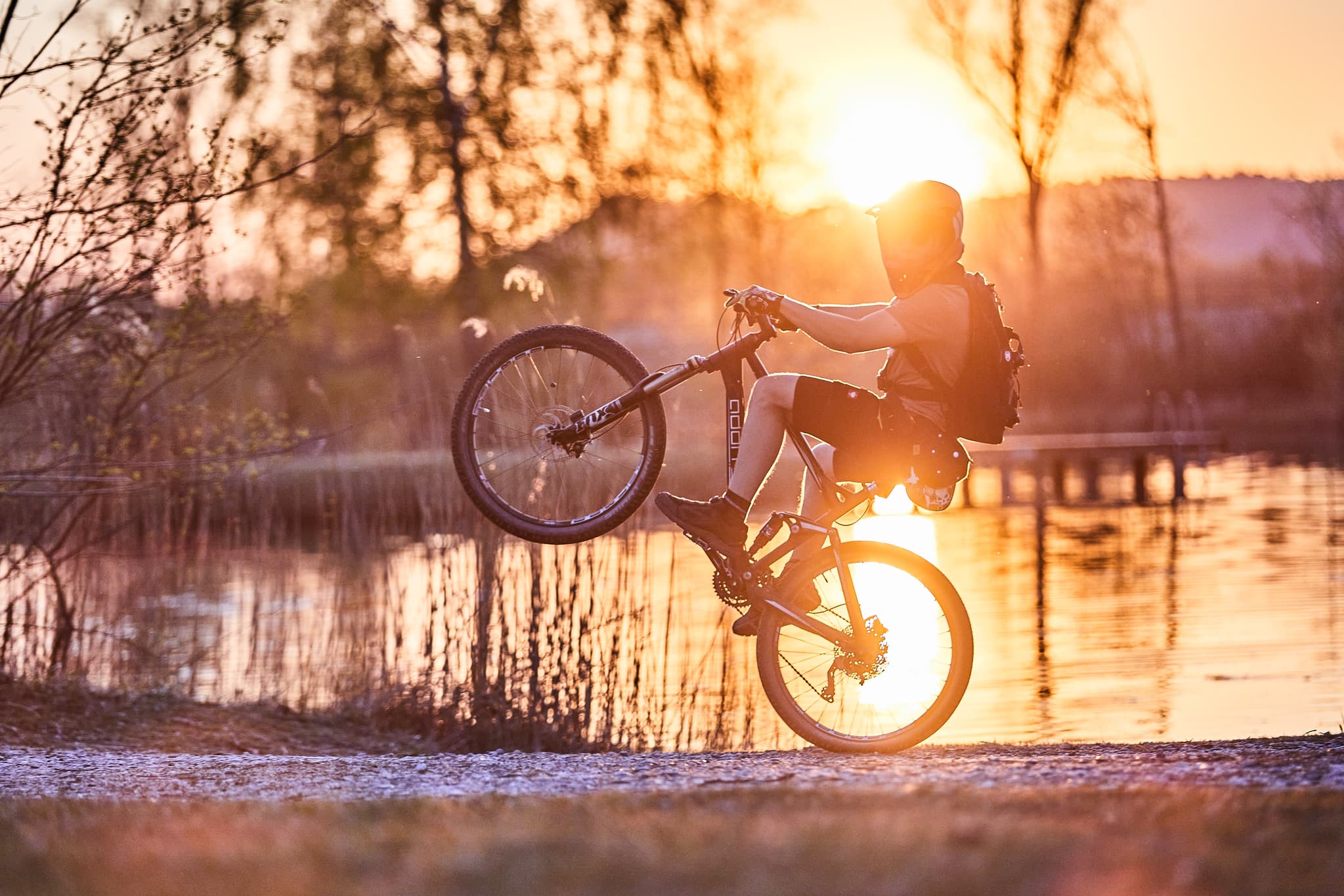 Ein Mountainbiker macht einen Wheelie am Ufer des Badesees in Perach bei Sonnenuntergang. Landkreis Altötting, Oberbayern, Deutschland, Region Inn-Salzach.