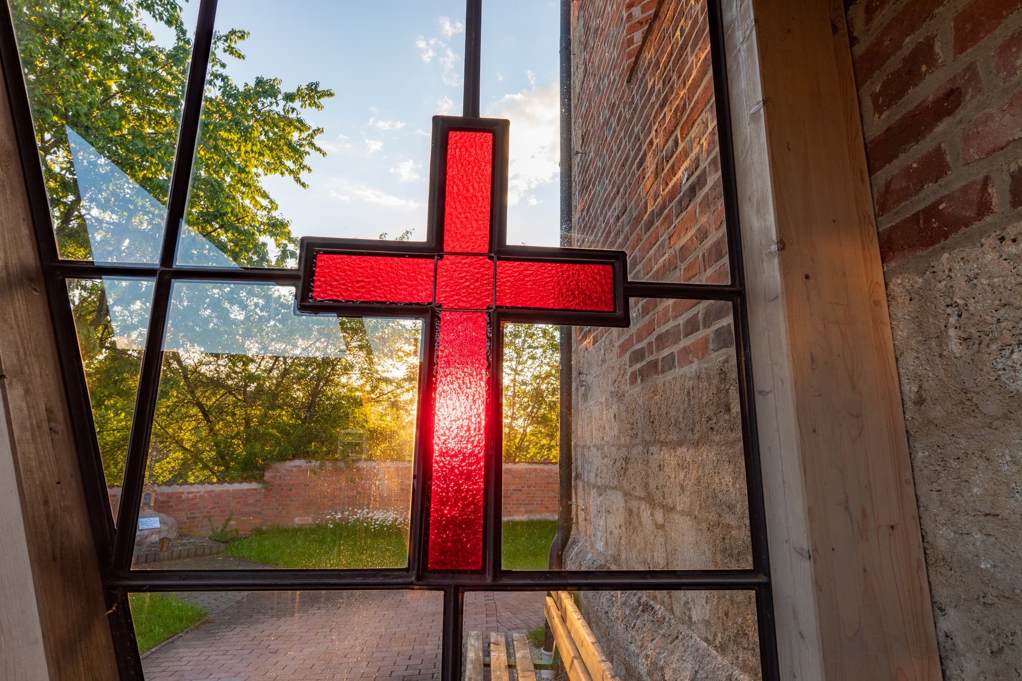 Kirchenfenster, rotes Kreuz. Blick auf Kirche Gumpersdorf, Zeilarn, Rottal-Inn, Niederbayern. Holzland, Deutschland. Mauerwerk, Grünflächen, Weg im Sonnenlicht.