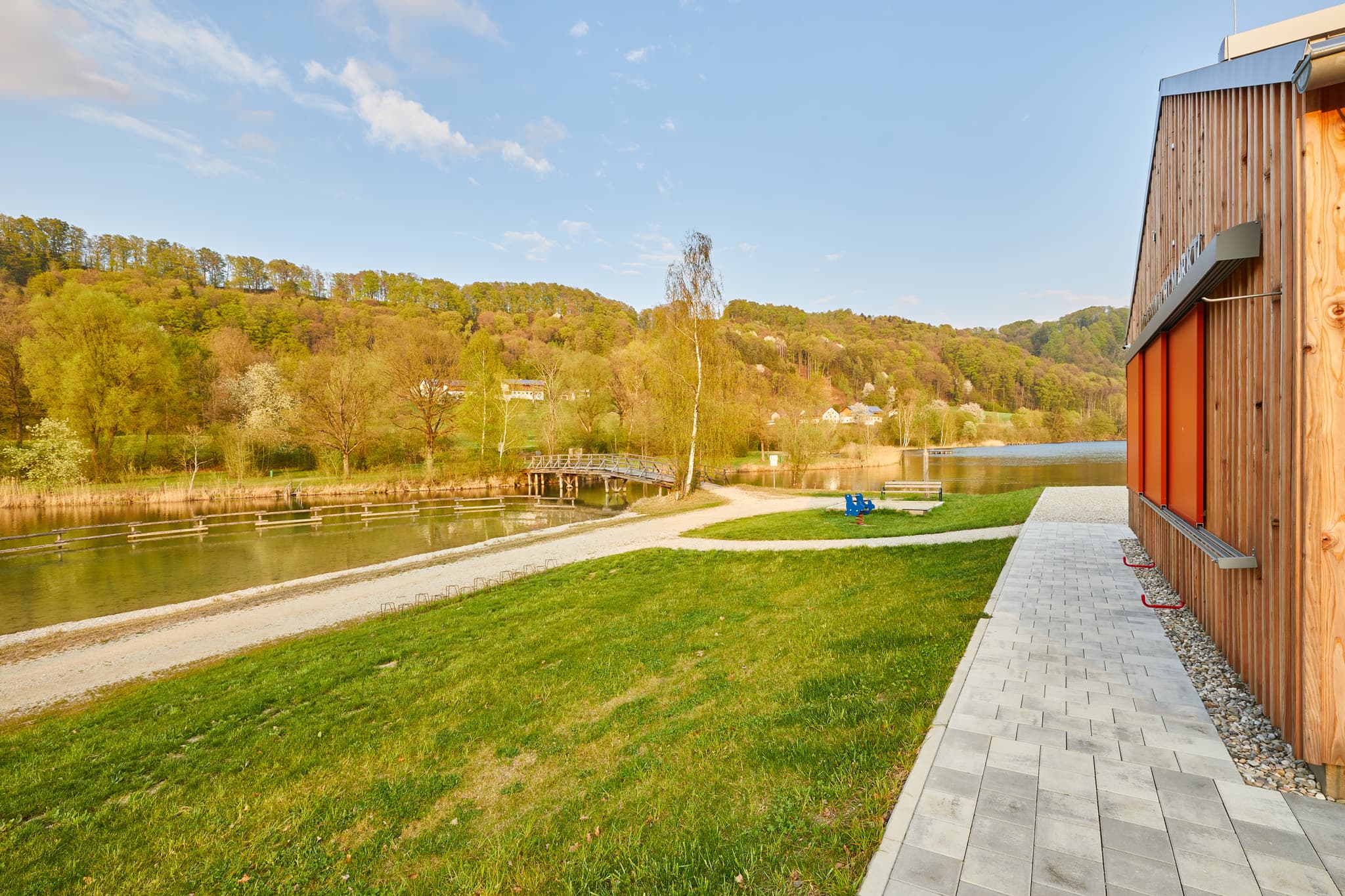 Badesee mit Kiosk und Umkleide in Marktl am Inn, Oberbayern, Region Inn-Salzach, Bayern, Deutschland. Ausblick auf den See und die umliegende Landschaft.
