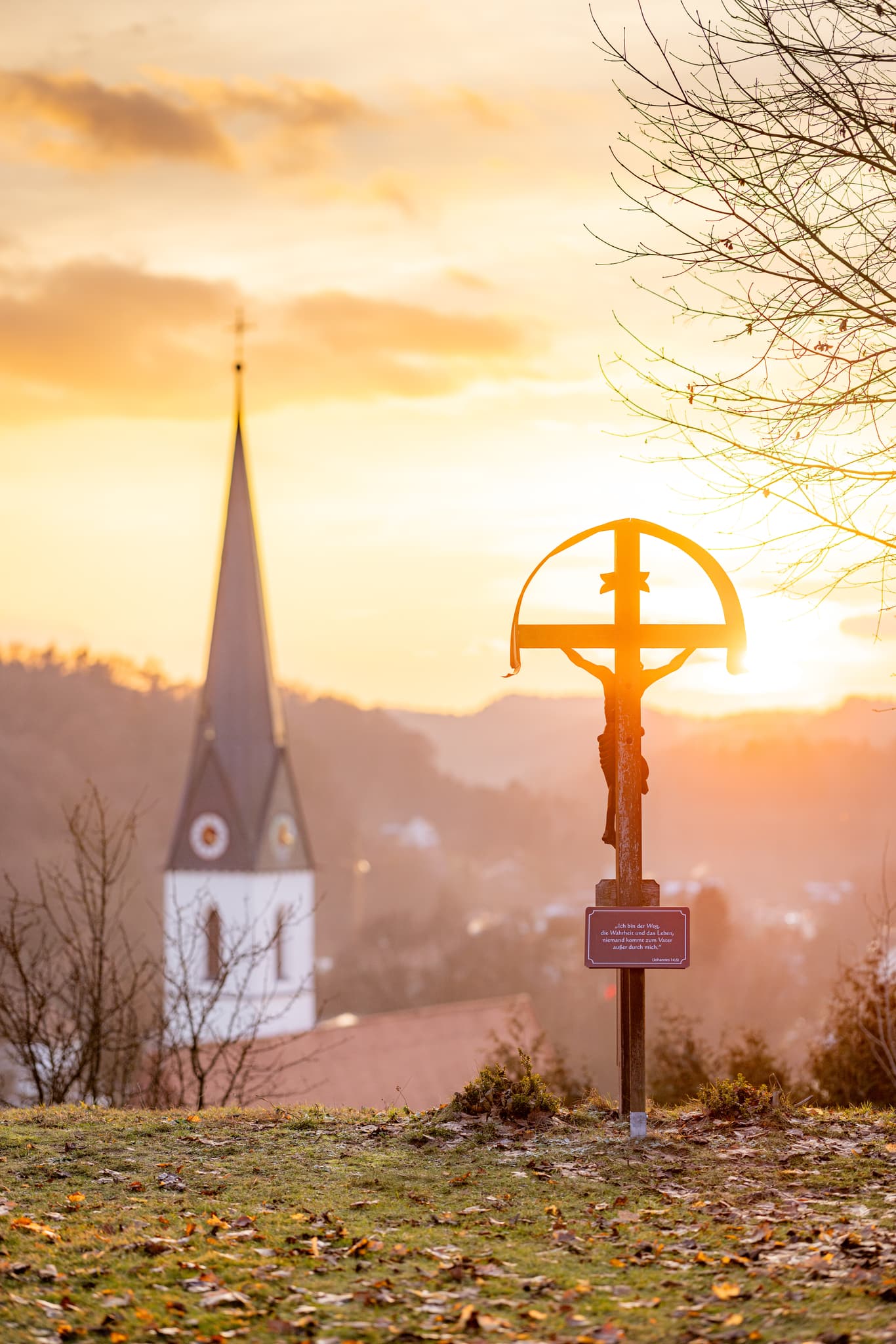 Wegkreuz am Zoglerberg in Reischach, Landkreis Altötting, Oberbayern, Deutschland. Abendstimmung in der Inn-Salzach Region. Mit Blick auf die Ortskirche.