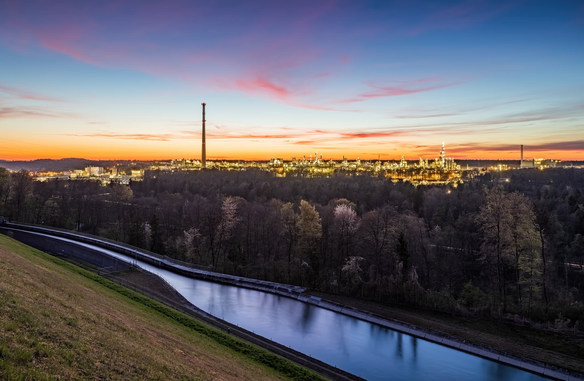 Industriepark Werk Gendorf mit Alzkanal, Burgkirchen, Landkreis Altötting, Oberbayern. Beleuchtete Anlage im Abendlicht unter farbigem Himmel, Inn-Salzach.