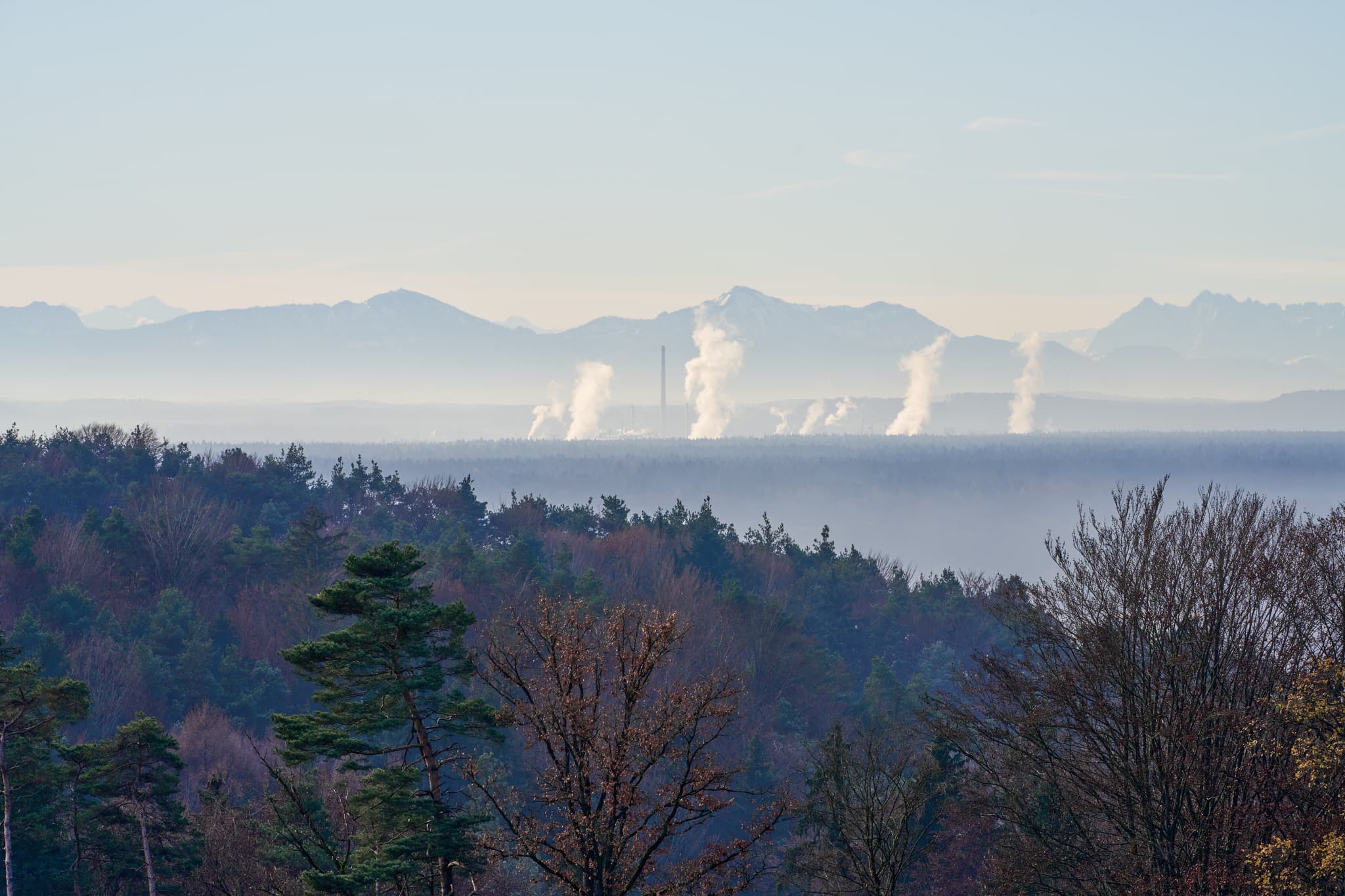 Blick vom Hasenberg, Perach, Altötting, Oberbayern. Zeigt Wälder, Industrieanlagen mit Rauch im Tal und Alpen im Hintergrund. Landschaft der Region Inn-Salzach.