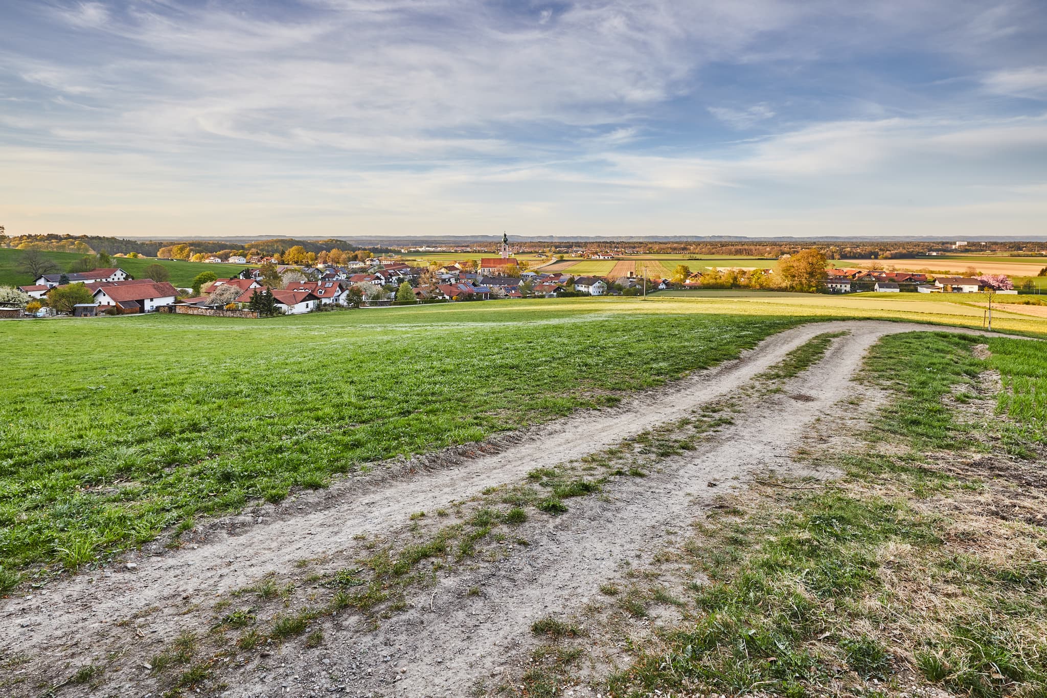 Ländliche Ansicht mit Feldweg durch grüne Wiesen. Im Hintergrund Dorf Mehring (Hörndlweg), Landkreis Altötting, Oberbayern. Teil der Region Inn-Salzach.