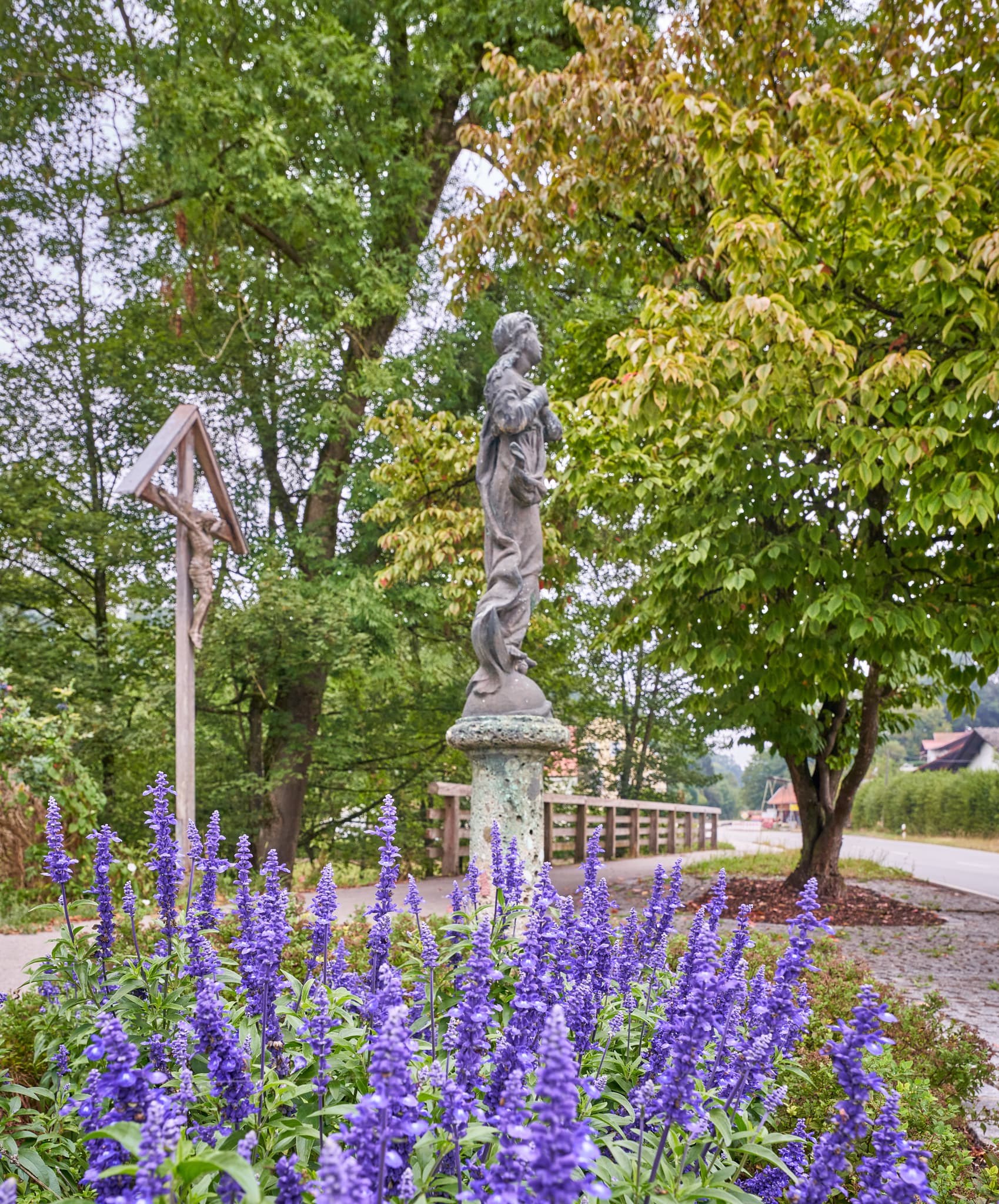 Wegkreuz und eine Statue mit lila Blumen im Vordergrund in Brandmühle, Reischach, Landkreis Altötting, Oberbayern, Region Inn-Salzach, Deutschland.