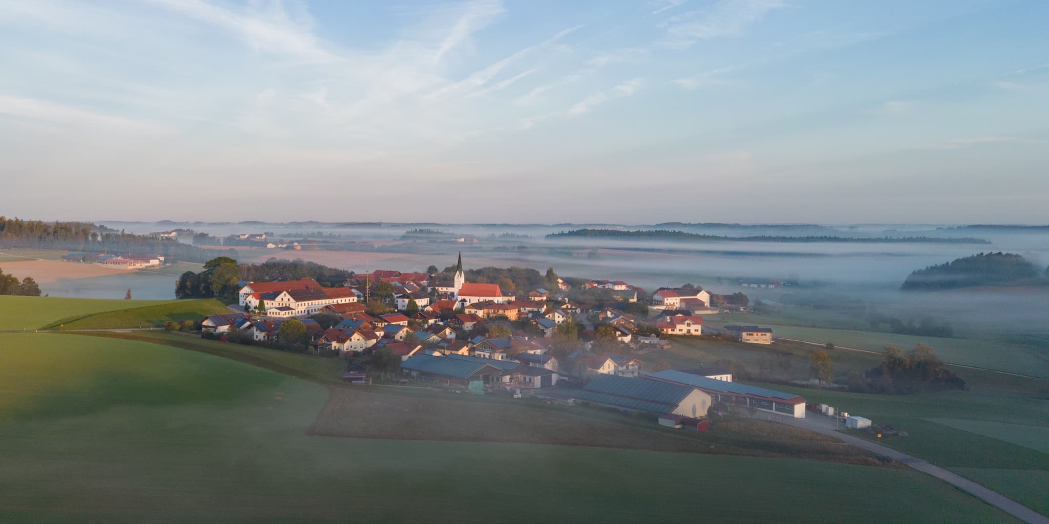 Luftbild von Arbing, Gemeinde Reischach, Landkreis Altötting, Oberbayern. Das Dorf im Holzland ist umgeben von Feldern und leichtem Morgennebel.