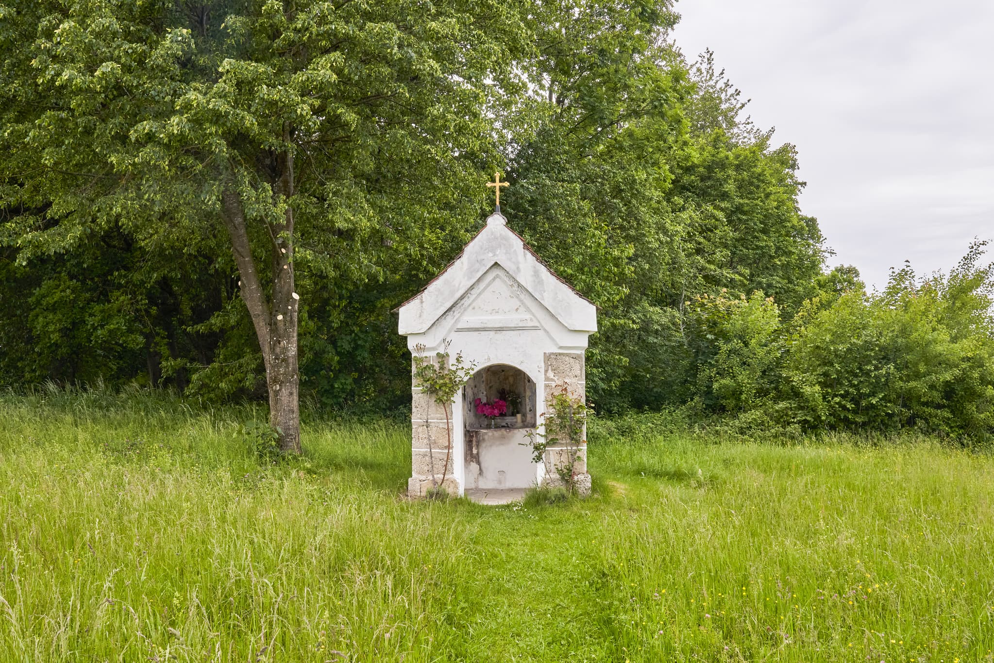 Bildstock am Weg in Höresham, Burgkirchen, Landkreis Altötting, Oberbayern. Ländliche Szenerie der Inn-Salzach Region mit Wiesen, Bäumen und Stromleitungen.