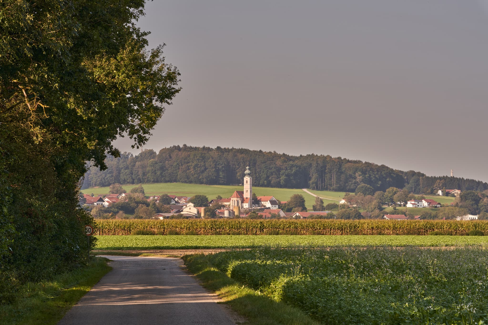 Blick auf den Pilgerweg bei Badhöring mit Dorf und Kirche im Hintergrund, umgeben von Feldern und Hügeln. Aufnahme in Mehring, Landkreis Altötting, Oberbayern