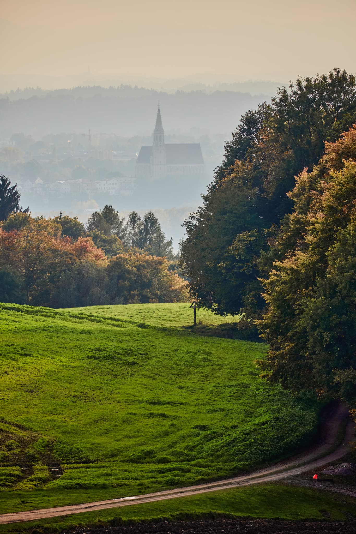 Blick von Friesing Aussicht in Reischach, Altötting, Oberbayern, Inn-Salzach, Deutschland. Zeigt hügelige Landschaft mit Kirche im Dunst, Feldern und Wegen.