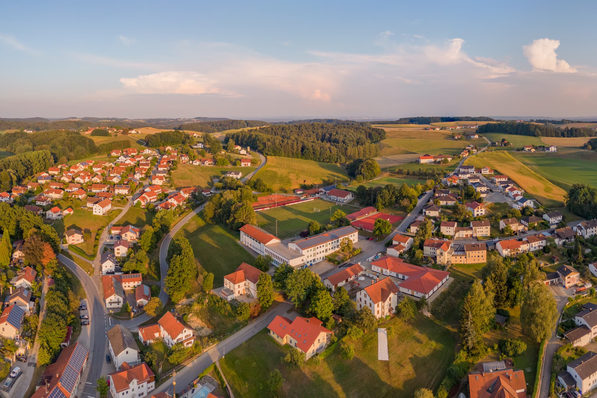 Luftbild von Reischach in Altötting, Oberbayern, Deutschland. Es zeigt die Schule und den Sportpatz, umgeben von Wohngebäuden und der weiten Landschaft.