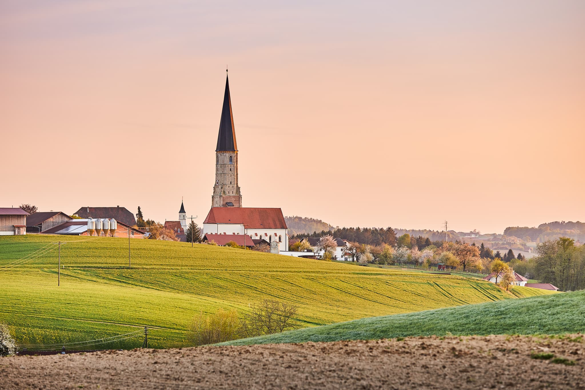 Kirche in Schildthurn, Ortsteil von Zeilarn, Landkreis Rottal-Inn, Niederbayern. Ländliche Landschaft des Holzlandes mit Hügeln und Feldern bei Dämmerung.