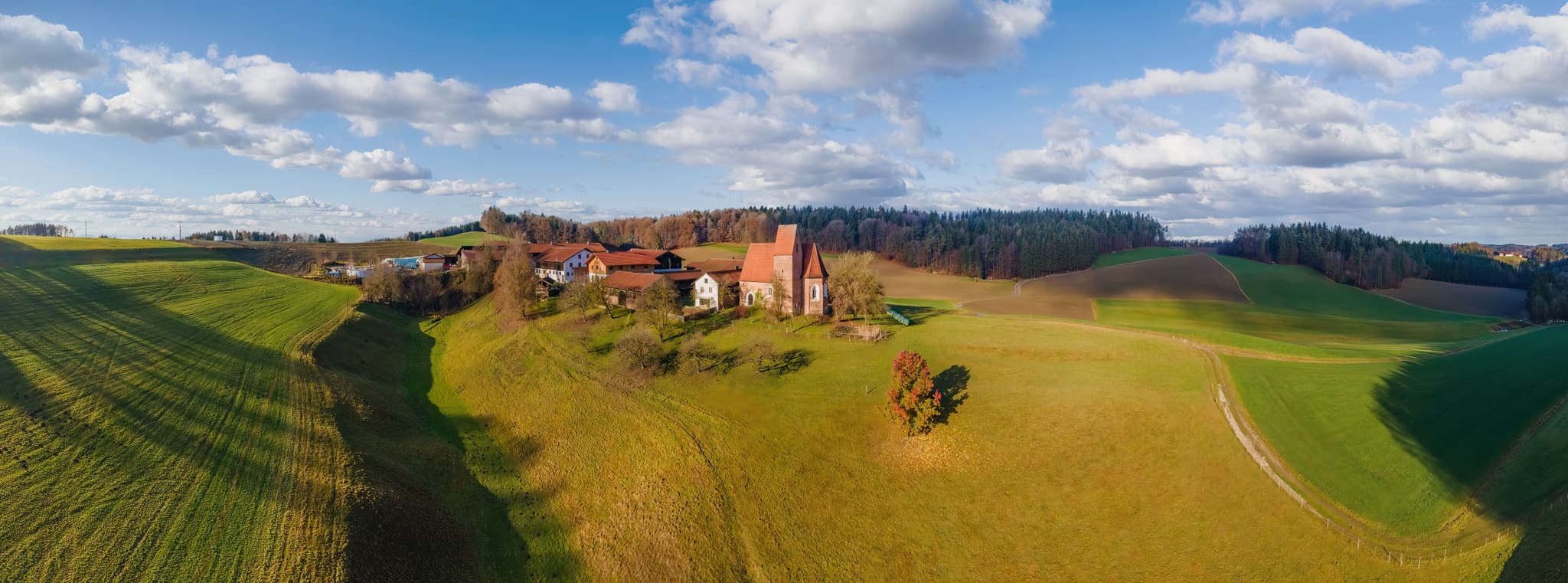 Berg Ortsteil mit Kirche St. Veit, Reischach, Altötting, Oberbayern. Die Inn-Salzach Region zeigt hügelige Felder und Wälder unter blauem Himmel mit Wolken.