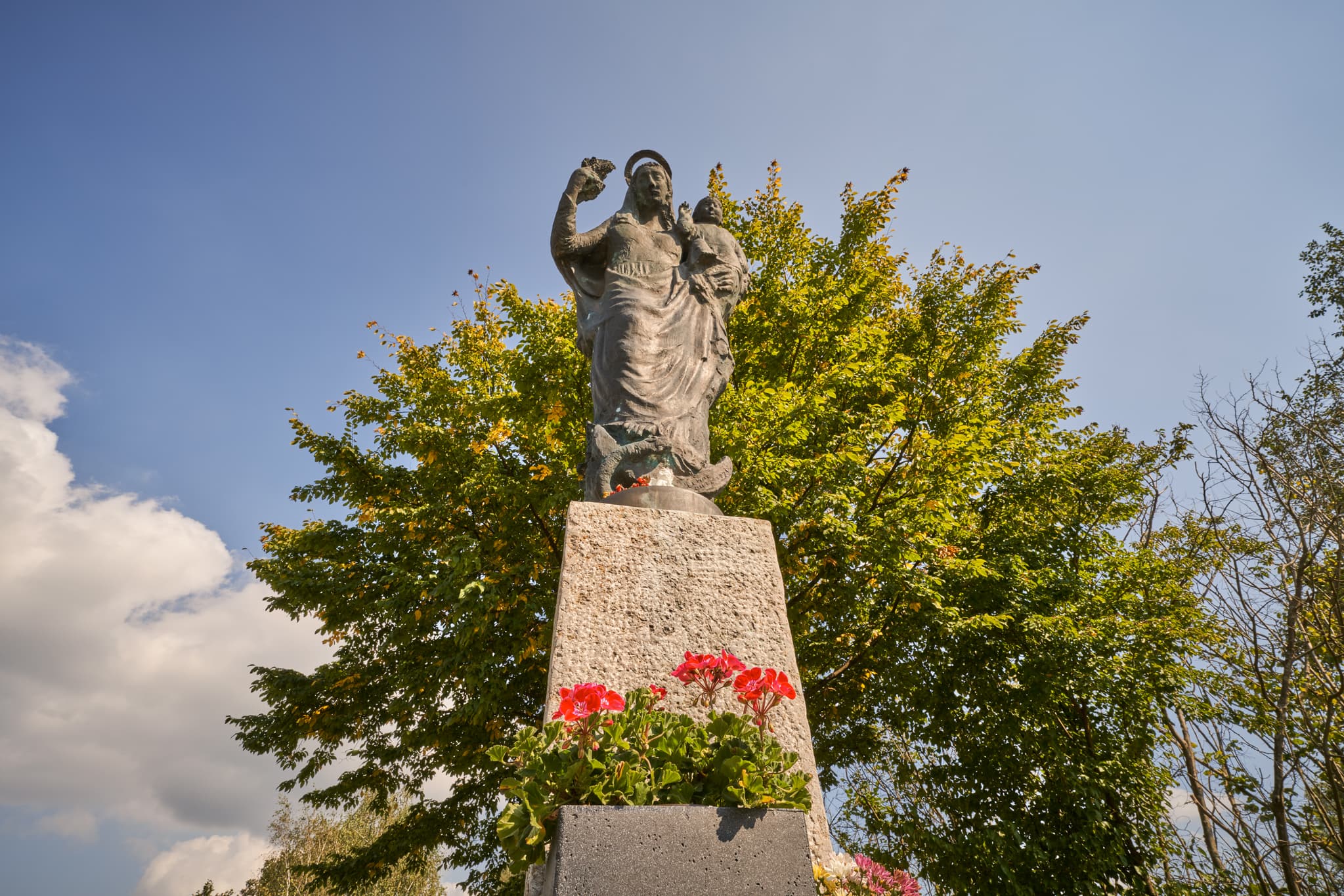 Maria mit Kind-Statue am B299 Parkplatz am Inn, Altötting, Oberbayern, Inn-Salzach, Deutschland. Unter blauem Himmel, Bäume rahmen ein.