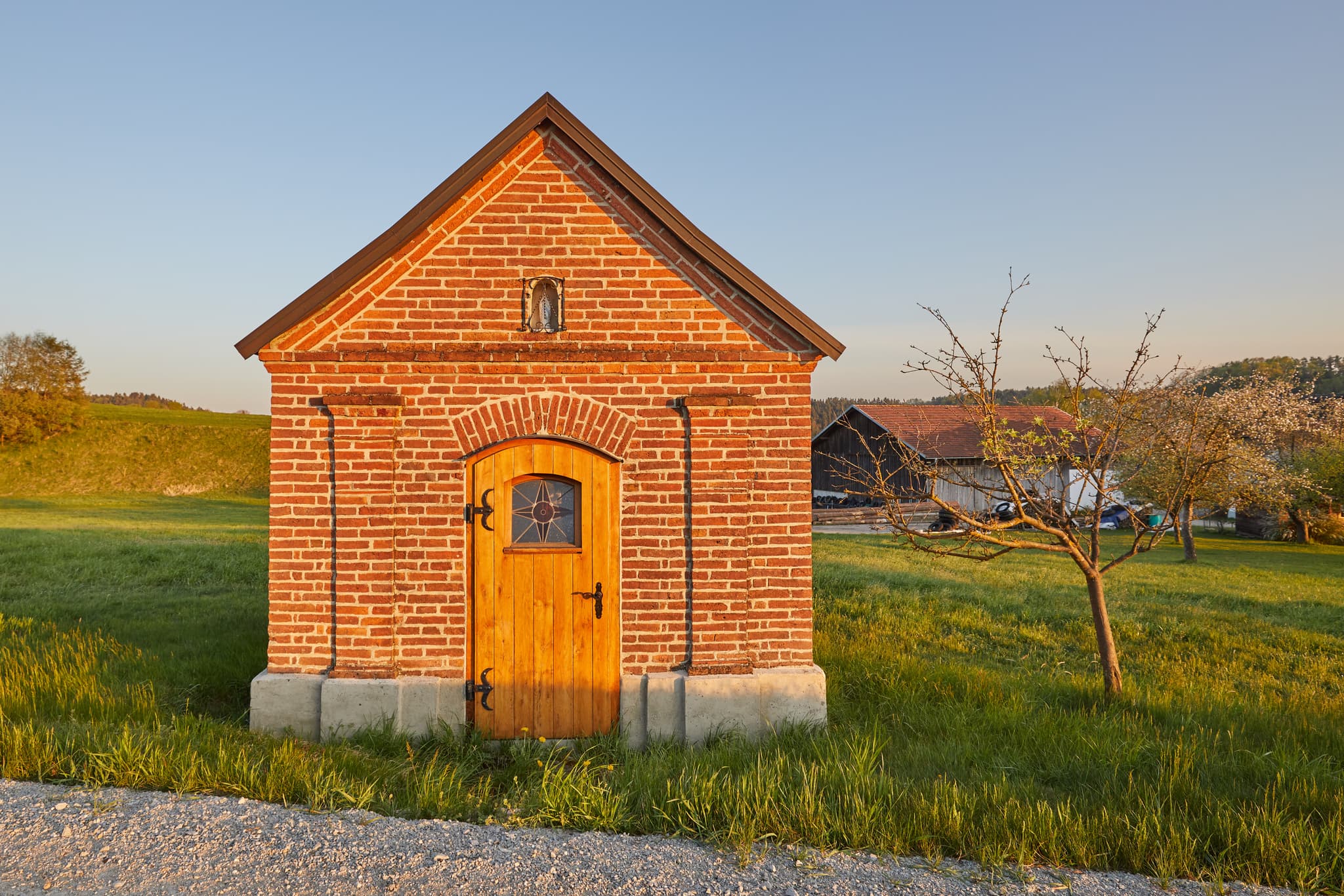 Kleine Backsteinkapelle im Abendlicht in Schildthurn bei Zeilarn, Landkreis Rottal-Inn, Niederbayern, Deutschland. Umgeben von grünen Wiesen und Bäumen.