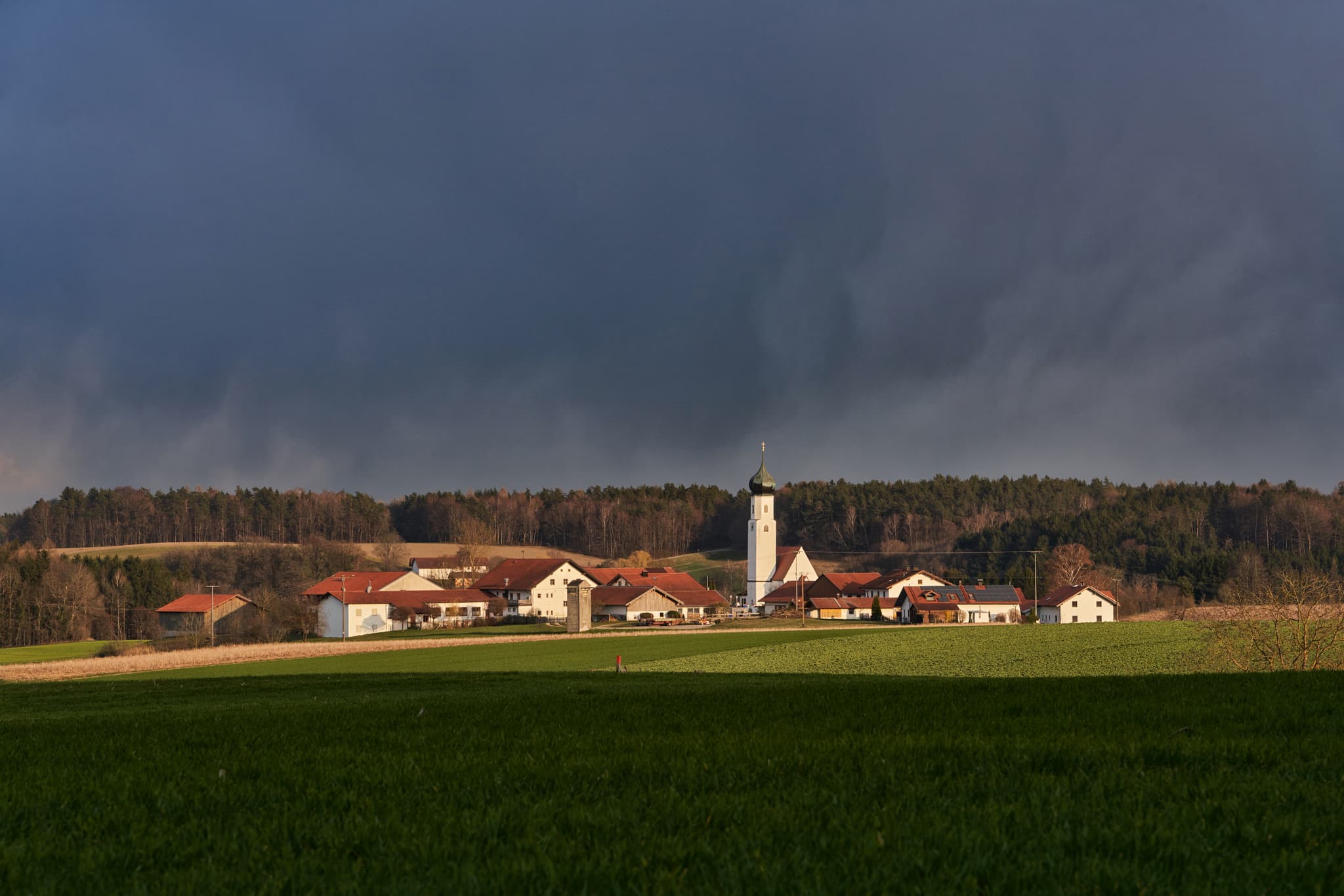 Ortsansicht Endlkirchen, Erlbach, Altötting, Oberbayern, Deutschland. Dorf mit Kirche, grüne Felder, Wald, dunkler Himmel. Typisch für die Inn-Salzach Region.