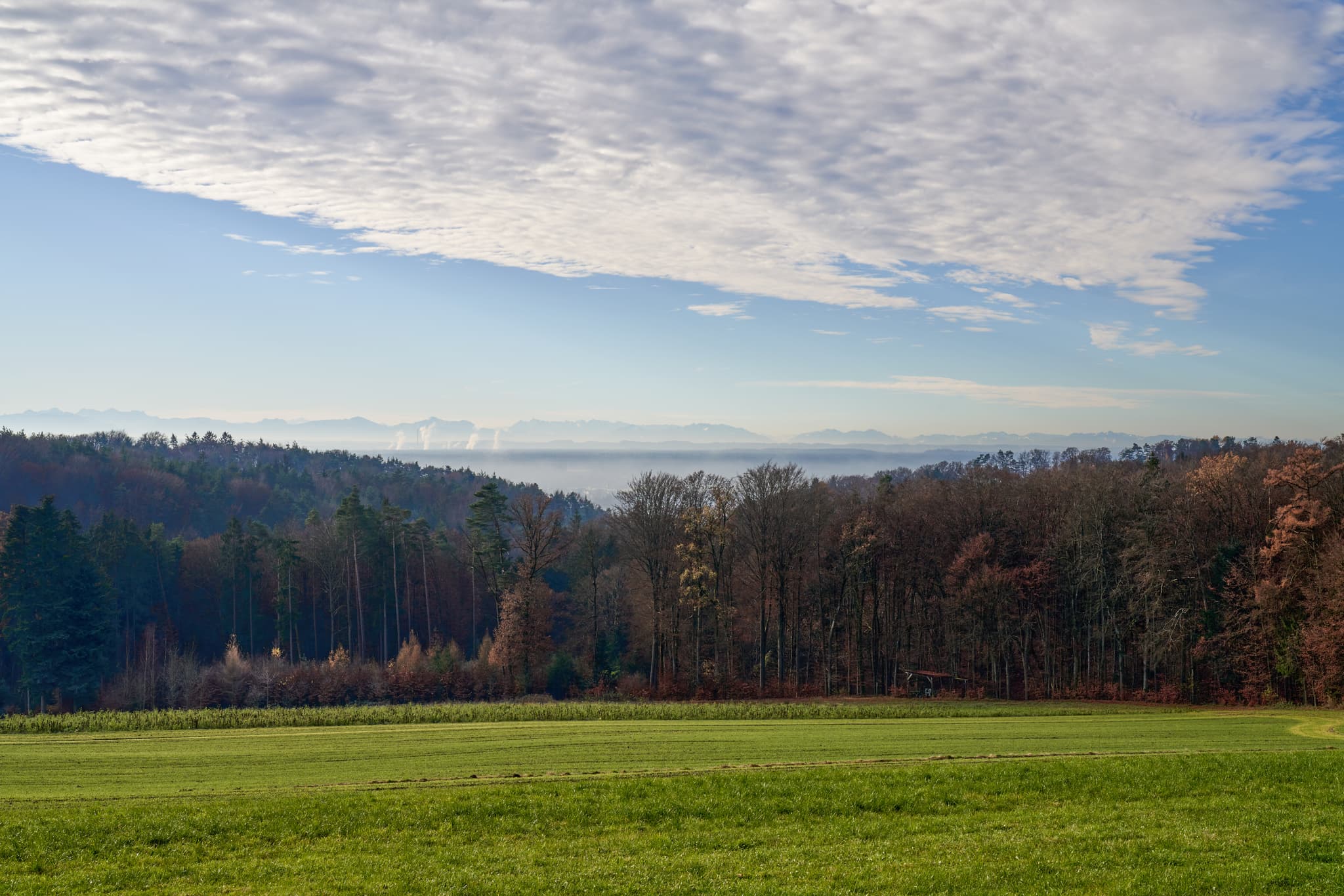 Blick vom Hasenberg, Perach, Altötting, Oberbayern. Zeigt Wälder, Industrieanlagen mit Rauch im Tal und Alpen im Hintergrund. Landschaft der Region Inn-Salzach.
