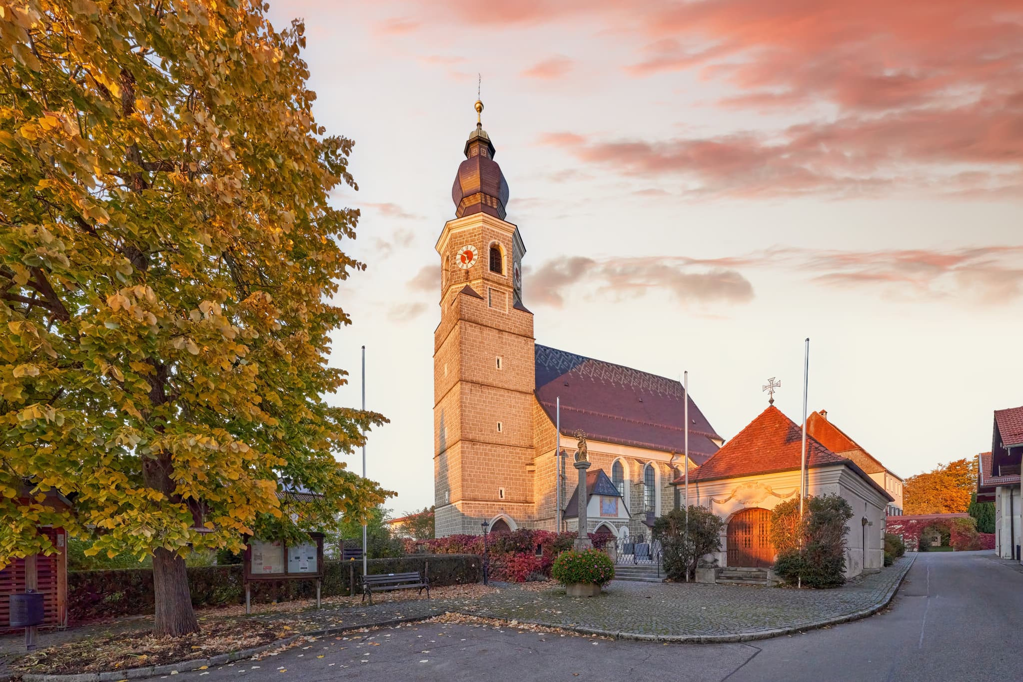 Kirche Mariä Himmelfahrt in Feichten, Altötting, Oberbayern, Inn-Salzach. Herbstlaub am Baum, rötlicher Himmel. Friedliche Szene.