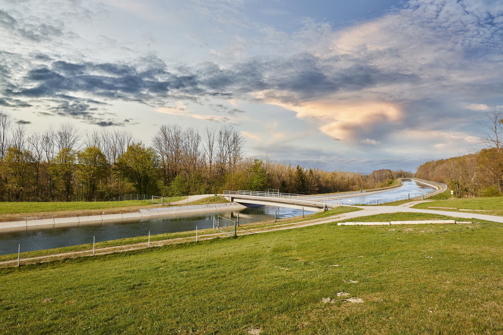 Der Alzkanal in Burgkirchen, Altötting, Oberbayern. Motiv: Kanal mit Brücke, grüne Wiesen, Bäume unter Wolkenhimmel.