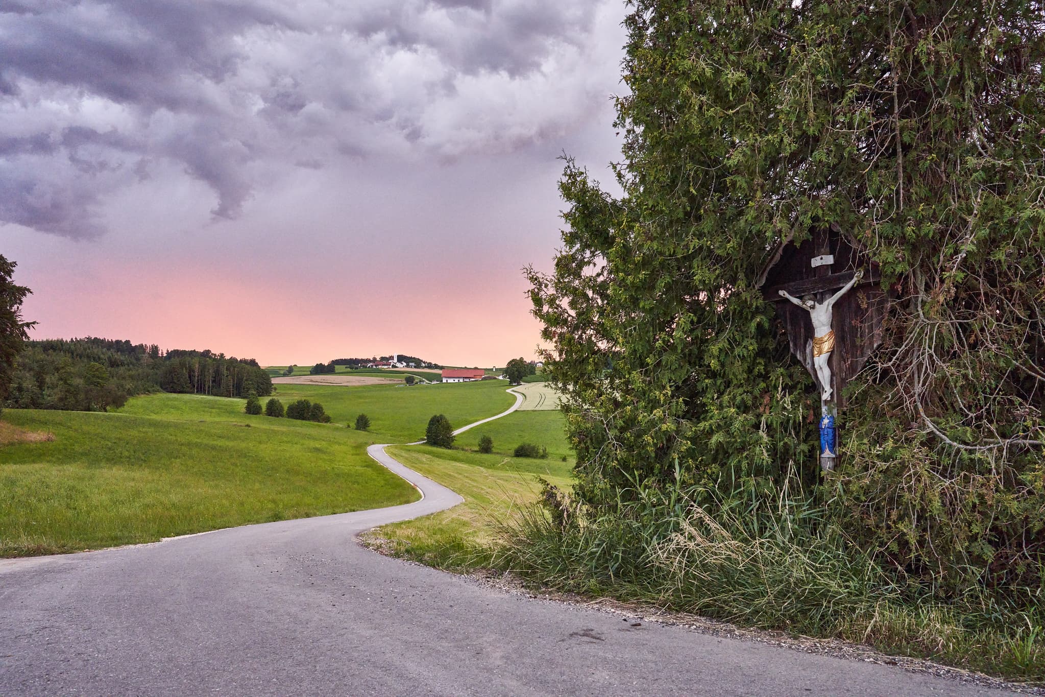 Arbing, Landkreis Altötting, Oberbayern, Deutschland. Ein Wegkreuz am Feldweg, umgeben von Wiesen unter dramatischem Himmel im Holzland.