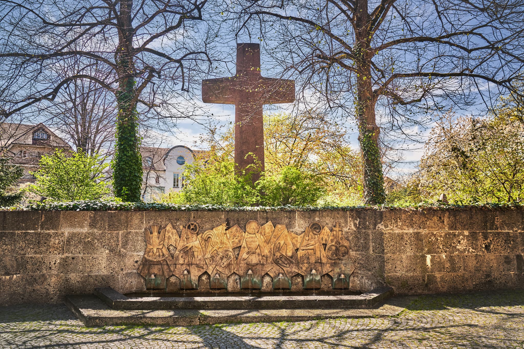 Kreuzweg mit Brunnen in Altötting, Oberbayern, Deutschland. Motiv zeigt kunstvollen Brunnen vor Steinmauer und Bäumen. Landkreis Altötting, Inn-Salzach Region.