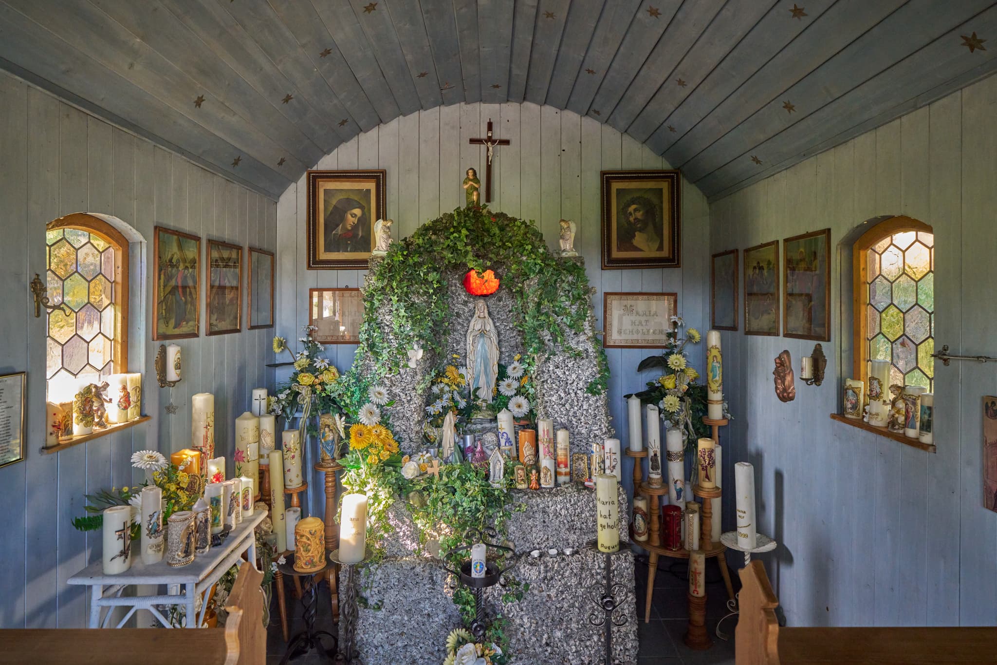 Innenansicht der Kapelle Stadlreith bei Tettenweis im Landkreis Passau, Niederbayern, Donau-Wald, Deutschland, zeigt Kerzen, Marienstatue und bunte Fenster.