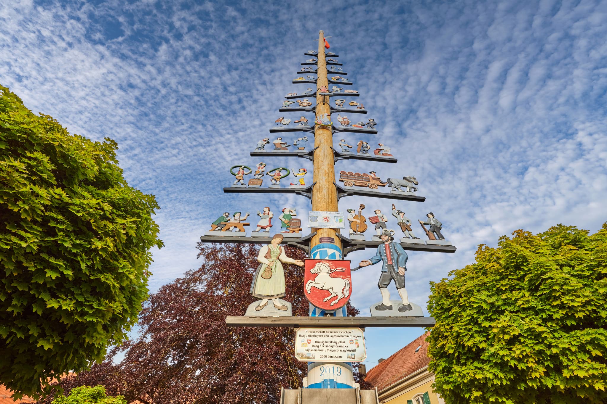 Ein festlich geschmückter Maibaum in Haag, Landkreis Mühldorf am Inn, Oberbayern, Region Inn-Salzach, Deutschland. Bei blauem Himmel und grünen Bäumen.