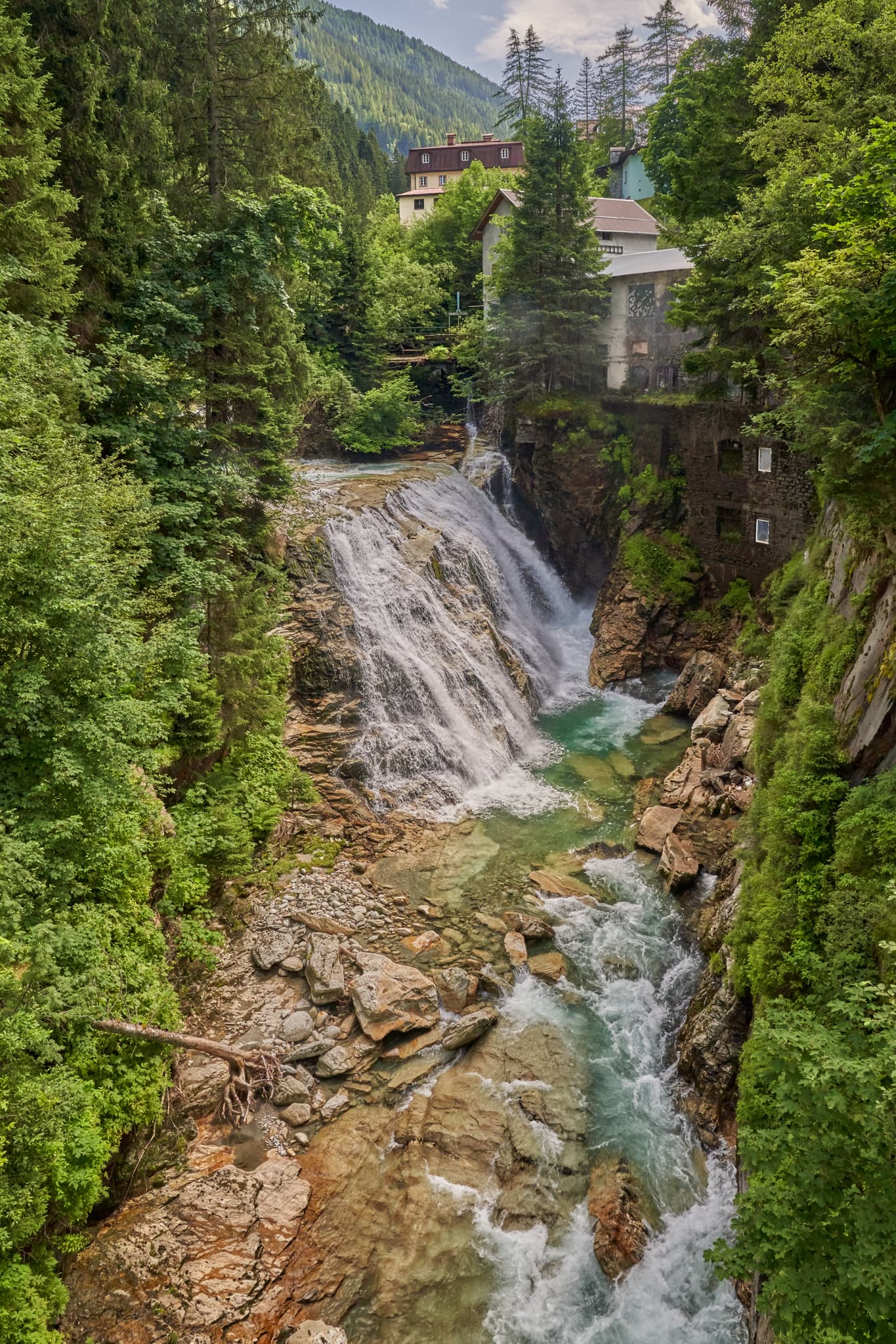 Wasserfall in Bad Gastein, St. Johann im Pongau, Salzburg, Österreich. Grüne Berglandschaft mit Wäldern, Felsen und Gebäuden prägt Kulisse.