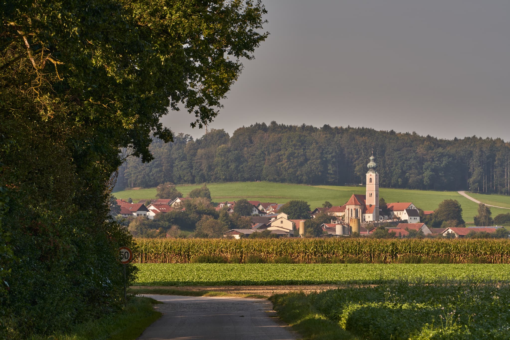Ansicht von Badhöring mit Kirche, umgeben von Feldern und Wald. Der Pilgerweg führt durch die Landschaft bei Mehring, Landkreis Altötting, Oberbayern.