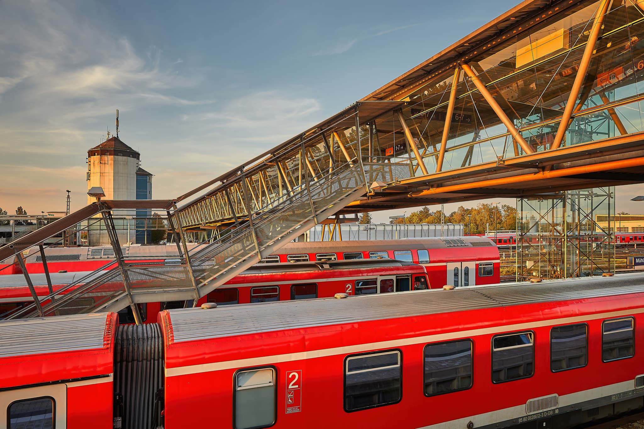Bahnhof Treppenabgang in Mühldorf am Inn, Oberbayern. Die Glasbrücke überspannt die Bahngleise mit roten Zügen. Ein Blick auf die regionale Bahninfrastruktur.