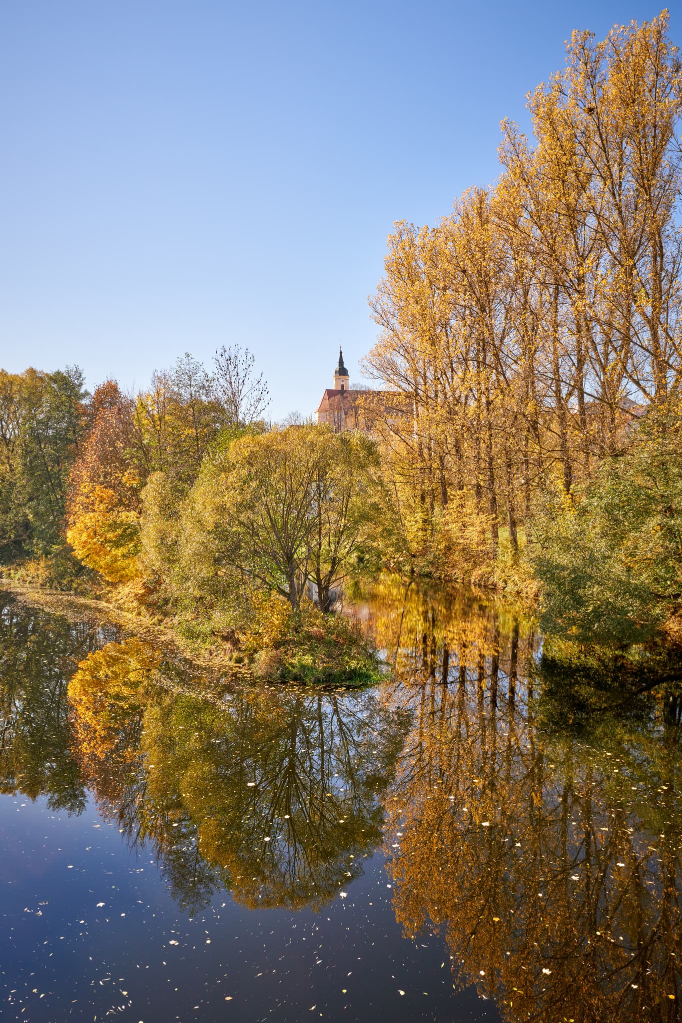 Herbstlandschaft am Schwarzen Regen bei Viechtach im Landkreis Regen, Niederbayern. Bäume spiegeln sich im Wasser, dahinter eine Kirche im Bayerischen Wald.