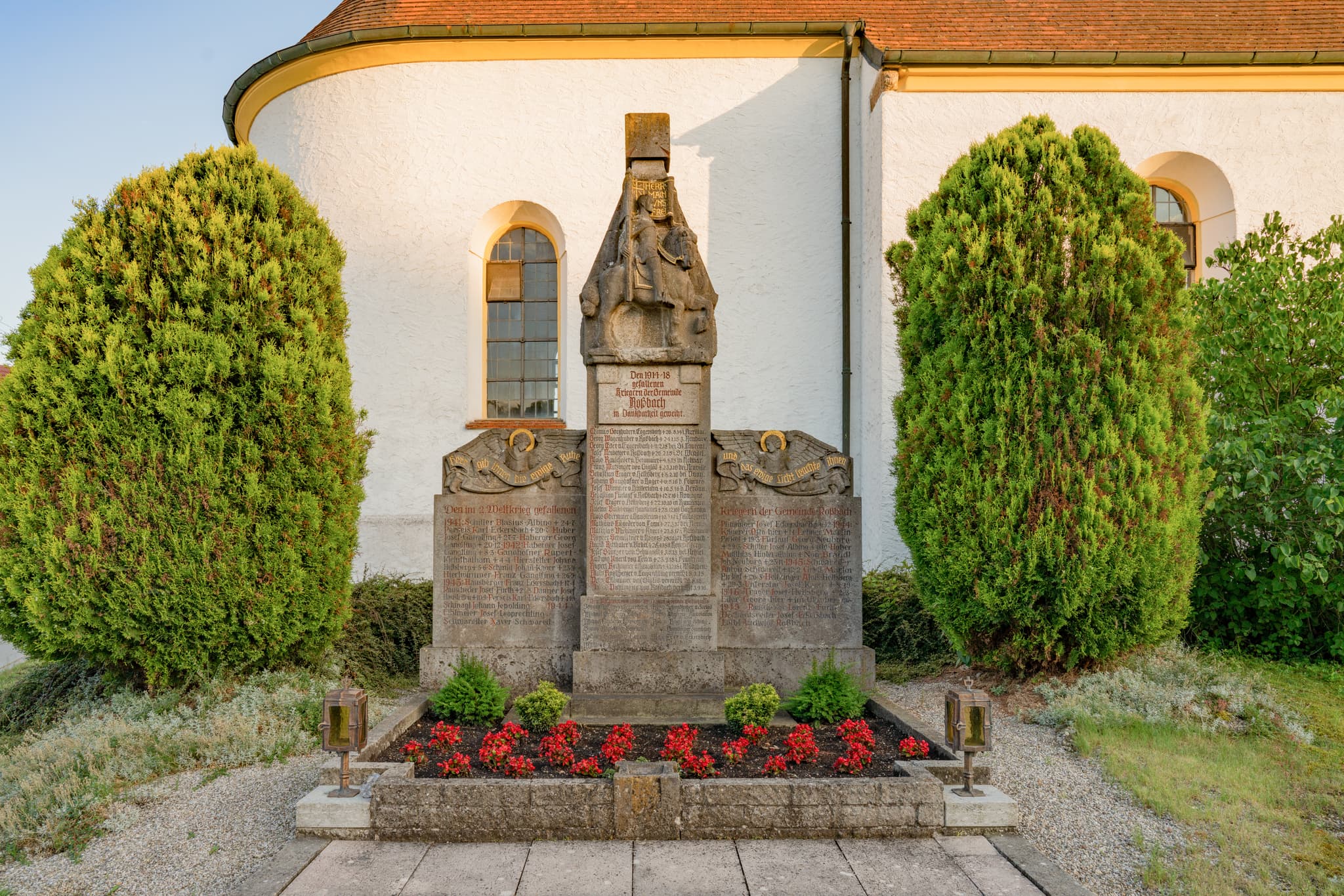 Kriegerdenkmal mit Büschen und roten Blumen vor Pfarrkirche St. Ägidius in Roßbach, Niedertaufkirchen, Mühldorf am Inn, Oberbayern, Inn-Salzach, Deutschland.