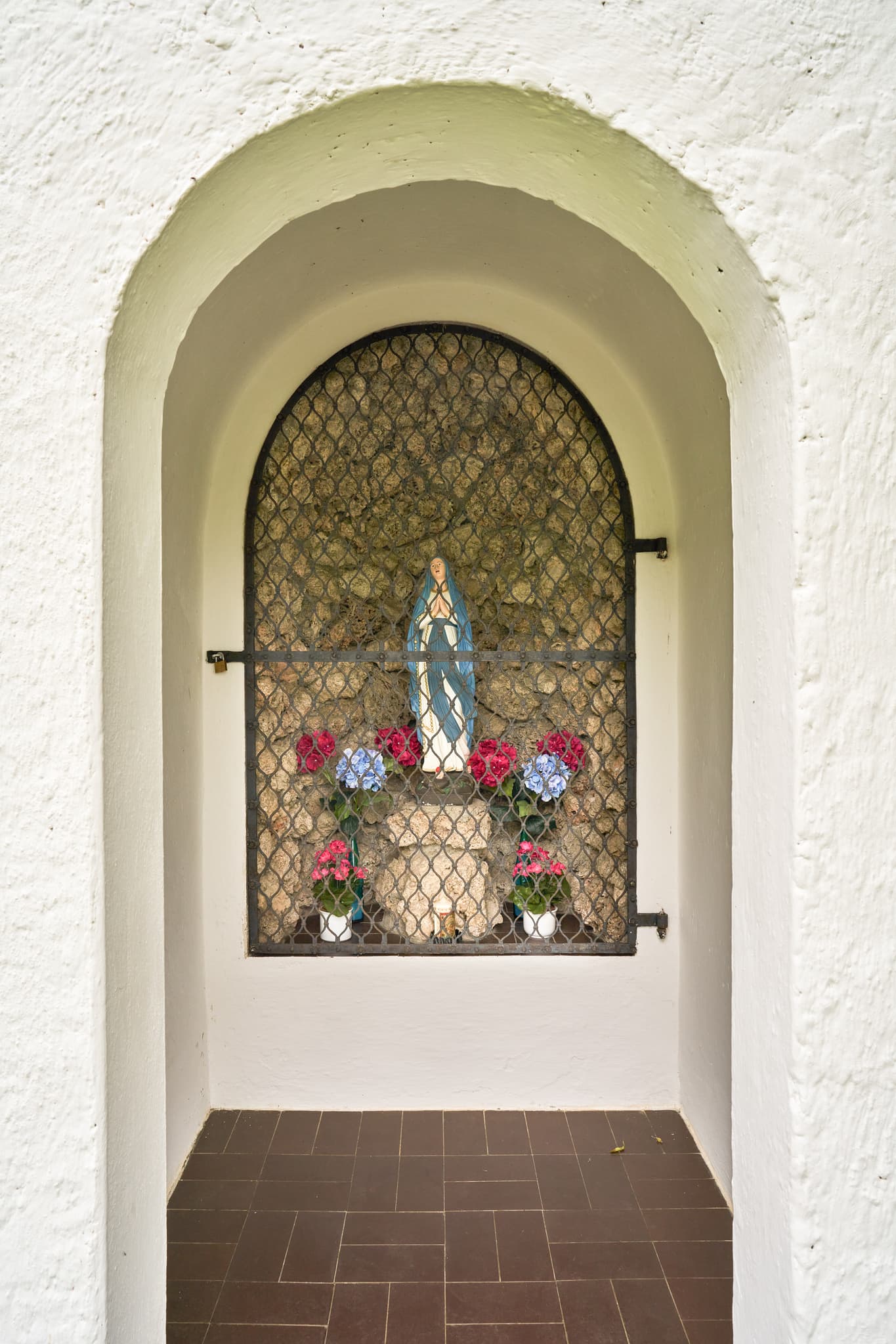 Ansicht einer Kapelle mit Marienstatue hinter Gitter und Blumen in Höresham, Burgkirchen. Im Landkreis Altötting, Oberbayern, Region Inn-Salzach, Deutschland.