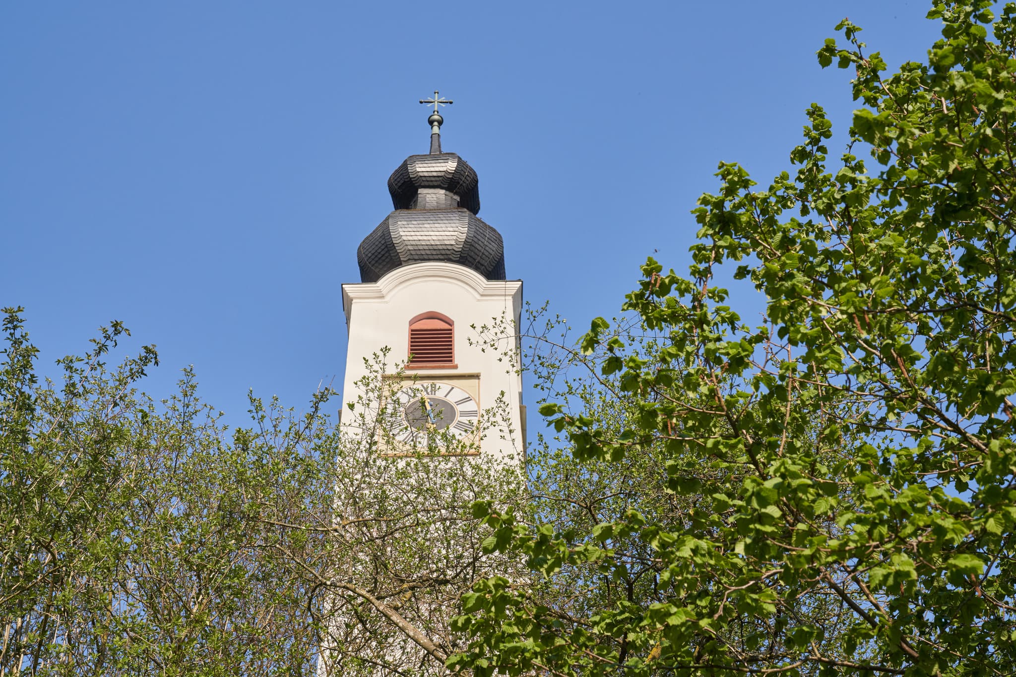 Kirchturm der Pfarrkirche Mariä Himmelfahrt in Perach, Landkreis Altötting, Oberbayern, Deutschland. Gotteshaus umgeben von Bäumen  der Region Inn-Salzach.