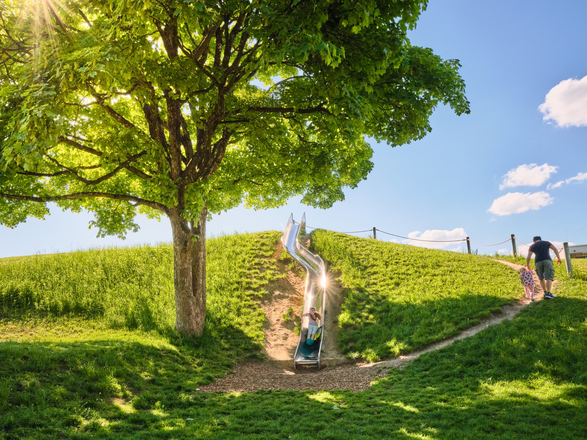 Kind auf Rutsche im Lindach Motorikpark, Burghausen, Altötting, Inn-Salzach, Oberbayern. Grüne Hügel mit Baum prägen die Szene in Deutschland.