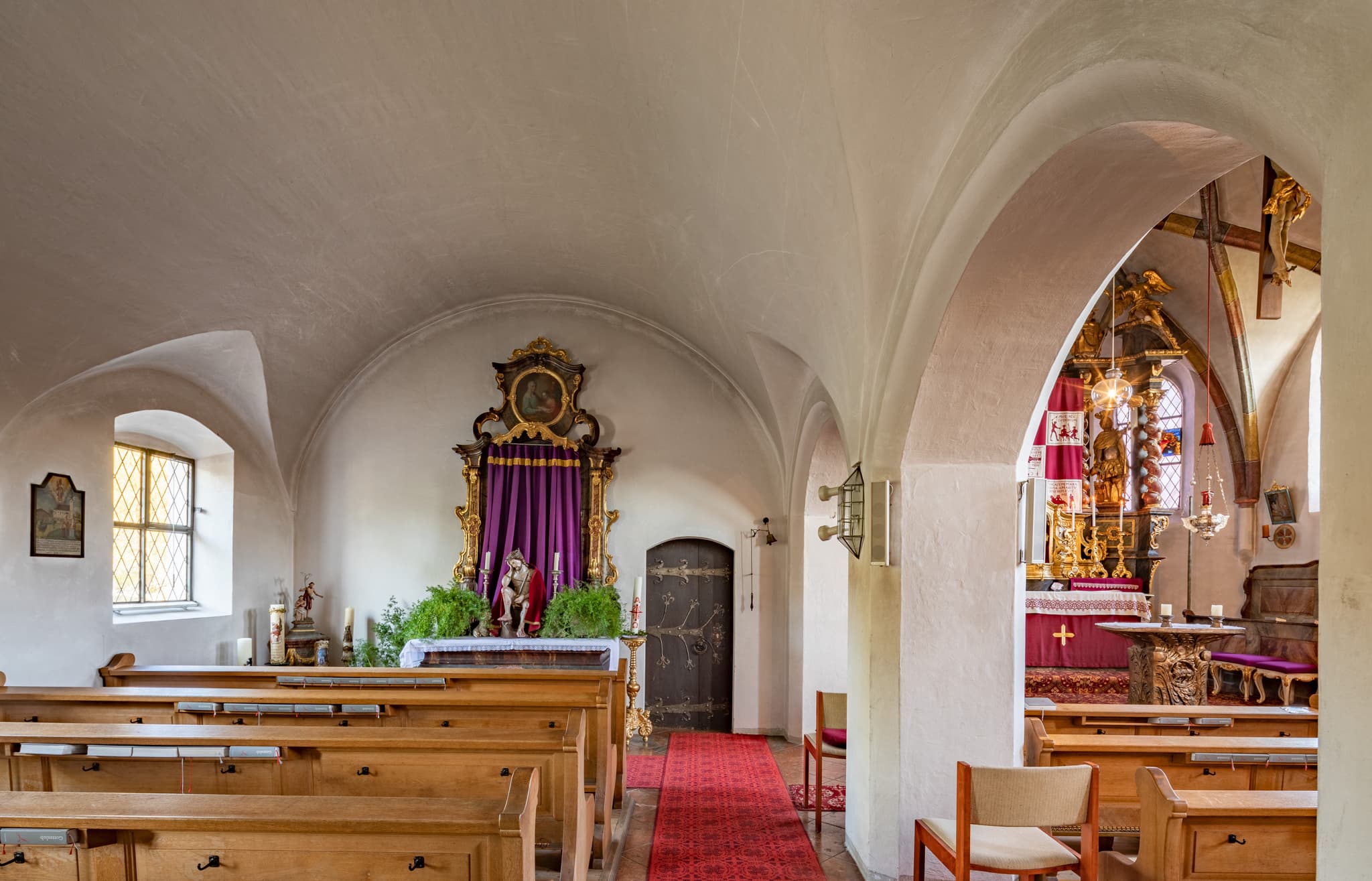 Innenansicht der Pfarrkirche Mariä Himmelfahrt in Perach, Altötting, Oberbayern. Blick  vom nebenraum auf das Kirchenschiff mit Holzbänken und Altar.